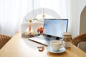 Laptop on a wooden desk by the window in an attic apartment. Gifts, advent candles, Christmas decoration and a coffee cup. Holiday