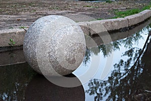 Large ball of granite in a puddle, in the park, on the background of asphalt and curb stone, in the reflection
