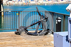 A large black anchor resting on a brown wooden dock surrounded by a black metal fence and vast deep blue ocean water