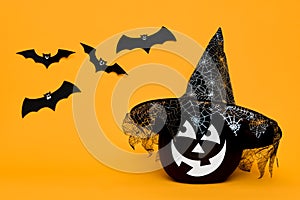 Large Black Halloween Pumpkin with cute smiling face wearing witch hat looking at flying bats over orange background.