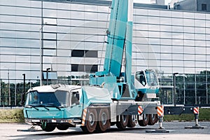 A large blue truck crane stands ready to operate on hydraulic supports on a platform next to a large modern building. The largest