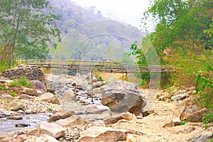 Large Boulder Rocks and river streams flowing in a mountain valley against hill, forest, and a Bamboo bridge in the background.