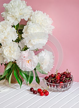 A large bouquet of peonies in a ceramic vase on the table, cherries in a bowl.