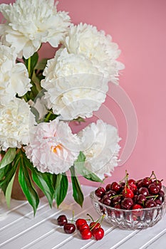A large bouquet of peonies in a ceramic vase on the table, cherries in a bowl.