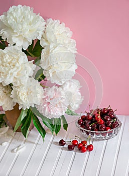 A large bouquet of peonies in a ceramic vase on the table, cherries in a bowl