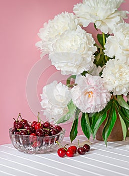 A large bouquet of peonies in a ceramic vase on the table, cherries in a bowl