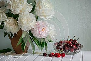 A large bouquet of peonies in a ceramic vase on the table, cherries in a bowl.