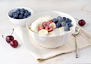 Large bowl of tasty and healthy oatmeal with fruits and berry for Breakfast, morning meal. Side view, white marble table