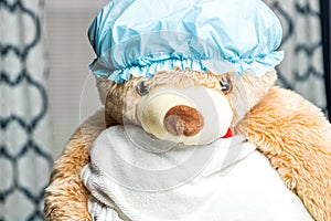 A large brown teddy bear in a shower cap and wrapped in a white towel