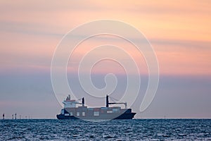 Large cargo ship with cranes carrying a load of shipping containers on the ocean at sunset.