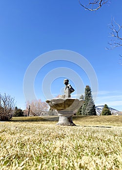 Large cement birdbath fountain & girl statue washing sits in a grassy yard with mountain & tree scenery beyond.