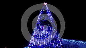 Large Christmas tree decorated with luminous garlands flashing on city square at snowy winter night. Camera flies around