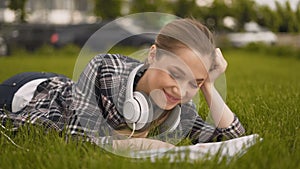 Large clan shot, a girl looks at the camera and smiles, big headphones, a student girl lies on the grass in the park and