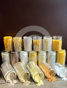 A large collection of various healthy grains and pasta in glass jars / containers, on a brown background.