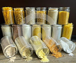 A large collection of various healthy grains and pasta in glass jars / containers, on a brown background.