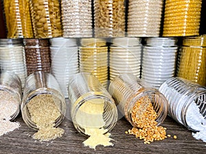 A large collection of various healthy grains and pasta in glass jars / containers, on a brown background.