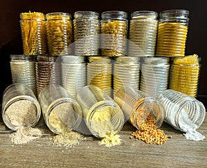 A large collection of various healthy grains and pasta in glass jars / containers, on a brown background.