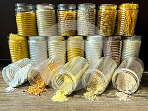 A large collection of various healthy grains and pasta in glass jars / containers, on a brown background.