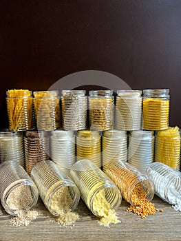 A large collection of various healthy grains and pasta in glass jars / containers, on a brown background.