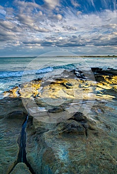 Large colourful rocks in calm shallow sea water soft waves blue sky with clouds, surfers in distance