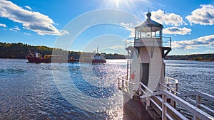 Large commercial ship on Maine river sails past small lighthouse with blue skies