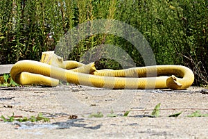 Large diameter bendable yellow plastic hose left on abandoned construction site surrounded with concrete and overgrown vegetation