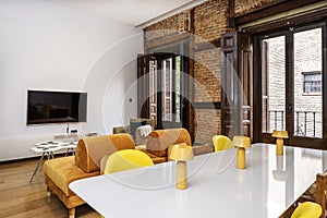 Large dining table in the foreground in a dining room with tv, large windows and exposed brick wall