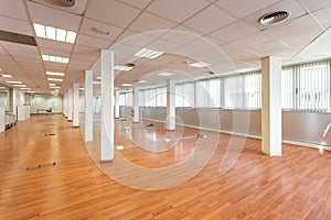 Large empty space with ceiling tiles, fluorescent lights, light brown laminate flooring and white painted columns. Large
