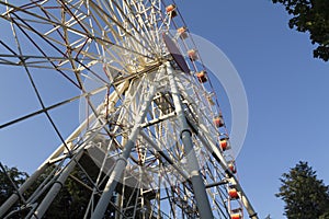 A large Ferris wheel in an amusement park with red and yellow cabins on a blue sky background. Trees in an amusement park