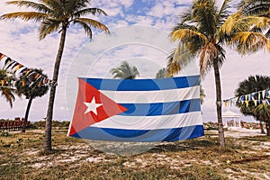 large flag of Cuba is strung between palm trees, on the beach of the Caribbean sea, against a cloudy sky. Beautiful tropical