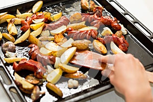 A large frying pan with fatty foods: sausages, potatoes in a rustic manner. Hand and wooden spatula