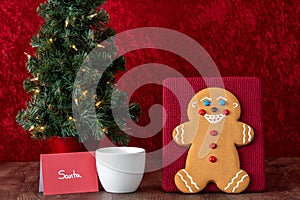 Large gingerbread cookie on a red background, Christmas tree with white lights, note to Santa, white mug with milk, wood table