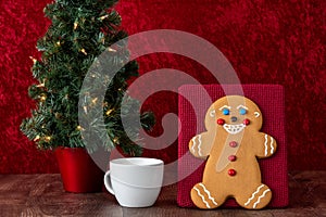 Large gingerbread cookie on a red background, Christmas tree with white lights, white mug with milk, on a wood table
