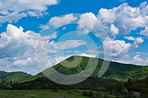 A large green hill with a pyramid shaped forest illuminated by the sun, and fluffy white clouds in the blue sky of a spring day