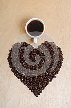 Large heart made of coffee beans and a cup of coffee on a light wooden background, top view