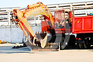 A large heavy yellow orange truck with a trailer, a dump truck and an excavator with a ladle are parked in a row at a construction