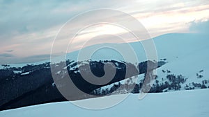 Large hills covered with a carpet of snow and fir forests against the backdrop of a cloudy sky and sunset