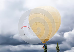 Large hot air balloon flying in the sky with clouds next to a motorized paraglider at an air festival.