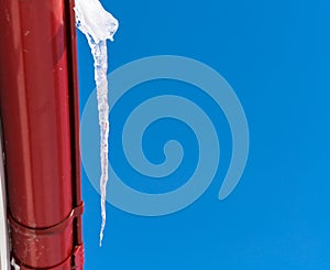 A large icicle against a clear blue sky hangs from a red drainpipe in winter on the eve of spring