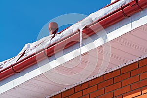 A large icicle against a clear blue sky hangs from a red drainpipe in winter on the eve of spring