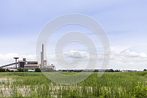 Large industrial factory building with conveyor and smokestack at water and green field on blue sky of bright day