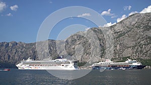 Large liner sails past a docked cruise ship off a mountainous coast