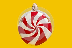 Large lollipop. Sweet gift. Christmas sweetness. Cute girl in shock holding a huge red and white candy on a yellow background.