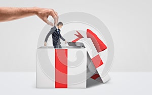 A large male hand getting a tiny businessman out from a white gift box with a red bow on white background.