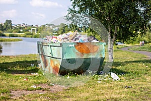 A large, metal, green and rusty container for garbage and household waste, standing in a Park near the lake