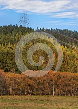 Large metal power pylon under electricity lines built in countryside with autumn grass and forest covered hills, blue sky
