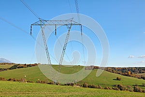Large metal power pylon under electricity lines built in countryside with green grass covered hills and small forests, blue sky