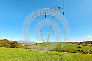 Large metal power pylon under electricity lines built in countryside with green grass covered hills and small forests, mount