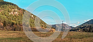 Large metal power pylons under electricity lines built in countryside with autumn grass and forest covered hills, blue sky