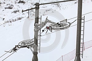 Large metal wheels for steel cables on top of ski lift supporting pillar, snow covered piste in background